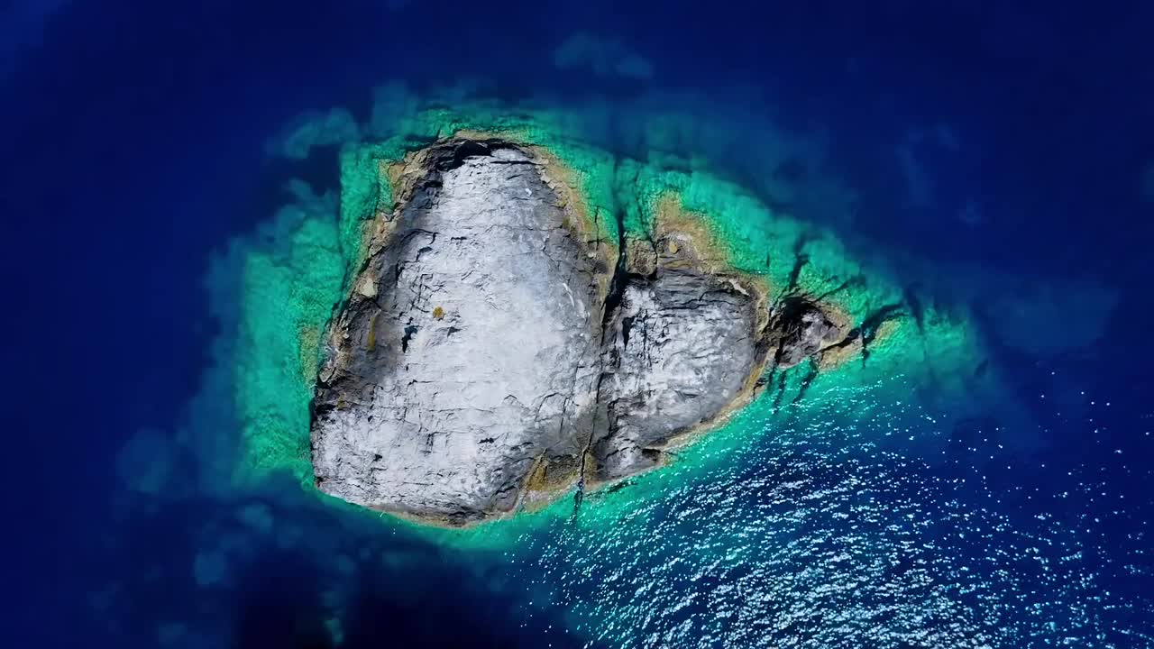 Vista aerea dall'alto con drone di un isolotto roccioso isolato circondato da un mare cristallino turchese che crea un sorprendente motivo naturale nelle acque dell'Egeo vicino alla Grecia.