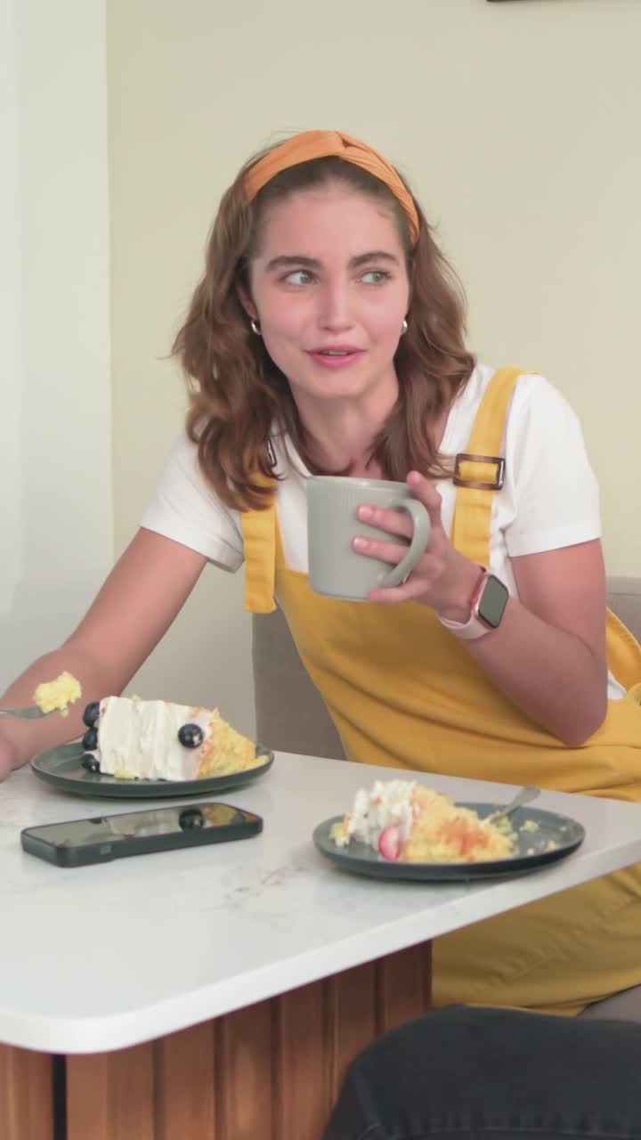 Video vertical: Mujer con vestido amarillo hablando mientras entra un amigo, con un pastel en un rincón y bebiendo de una taza gris.