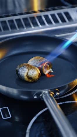 two pieces of fish rolled lie on the frying pan. chef uses a torch to burn fish skin scales. close up. vertical video.