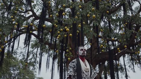 shaman with face painting standing under a tree decorated with fruits and black fibers, performing a sacred ritual in a natural setting, evoking spirituality and ancient traditions
