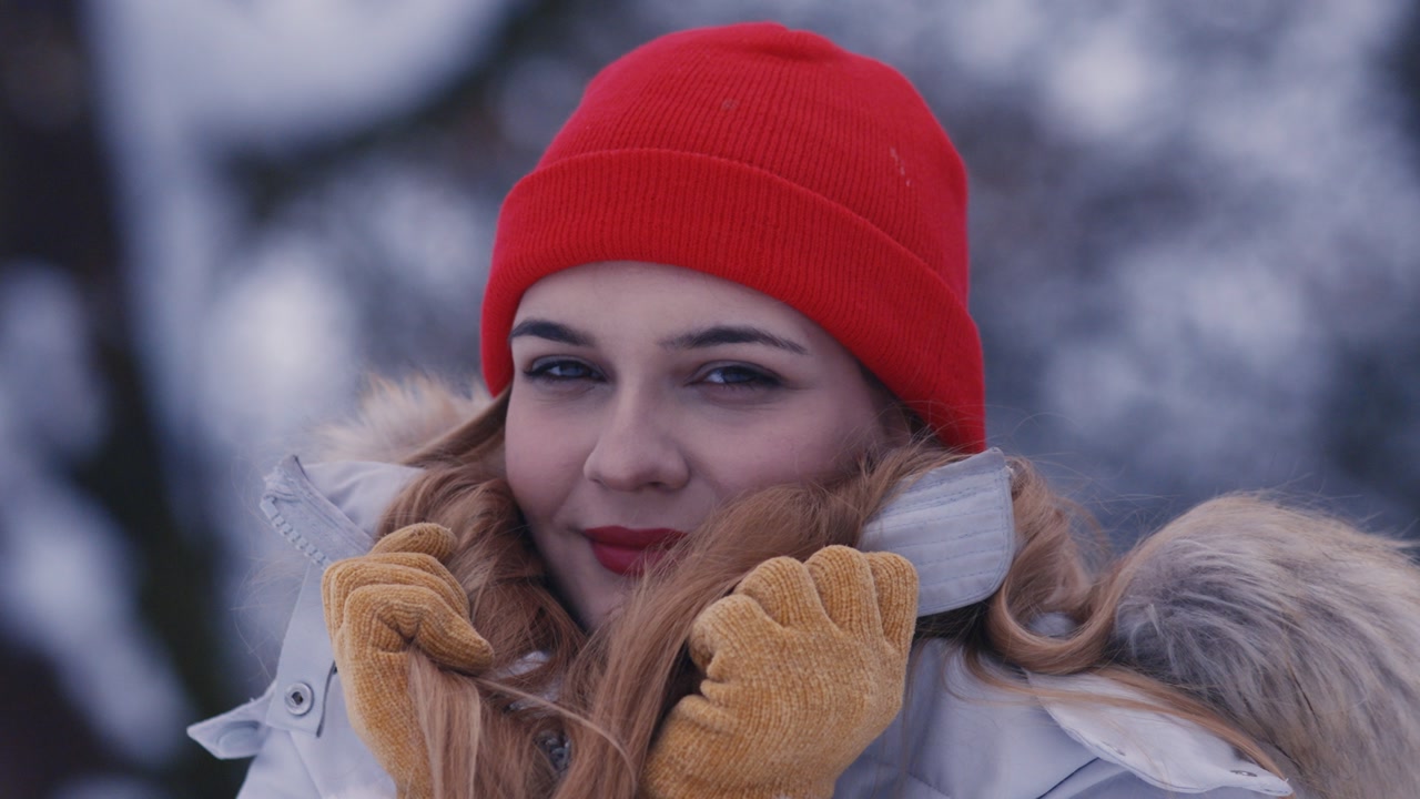 Close up portrait of a young woman wearing yellow gloves and a red beanie, pulling her jacket collar up in a snowy forest.