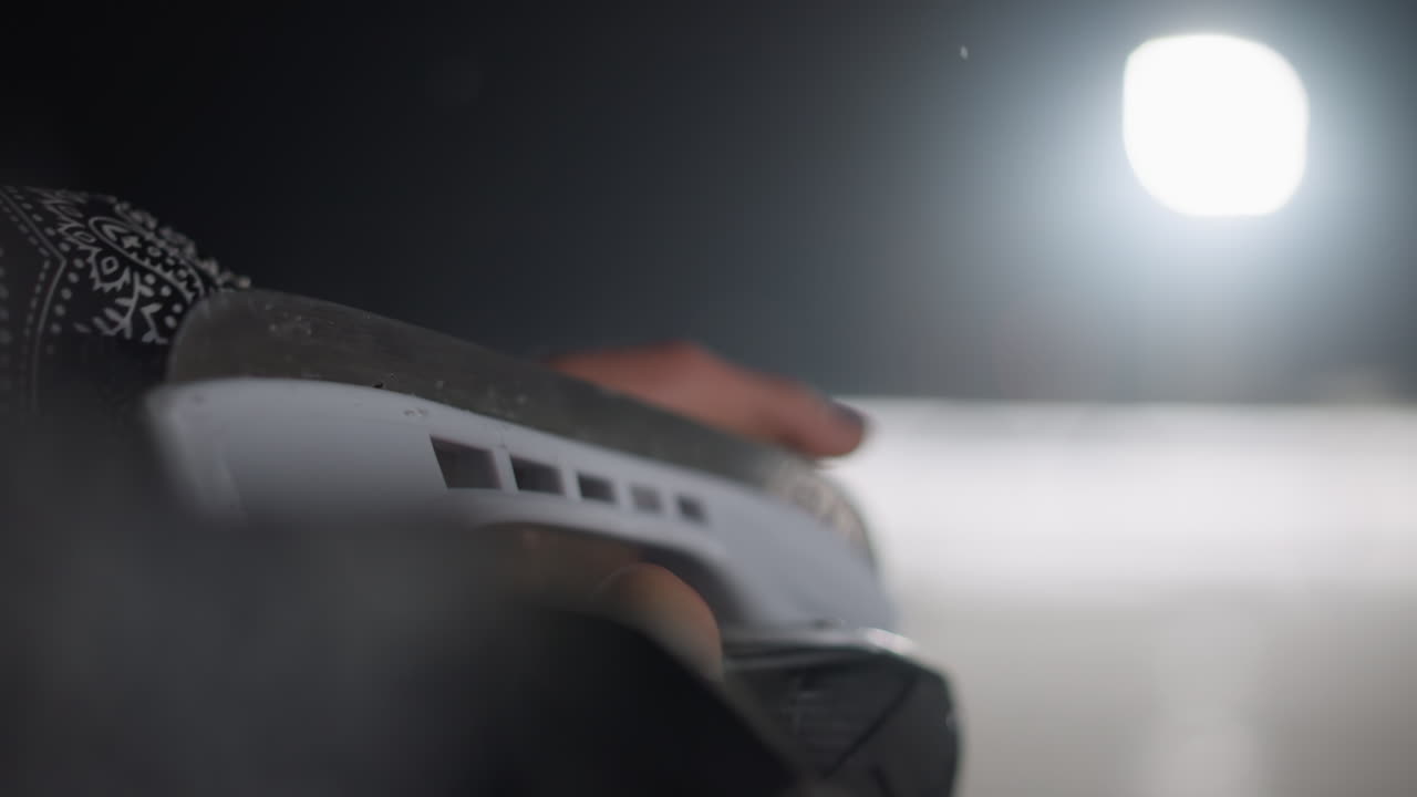 Close up rear view of skater seated gliding fingers across skate blade under bright night light with soft blur of snowy surface in background, evoking quiet focus and wintry atmosphere