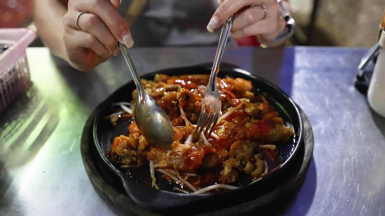 Hands use fork and spoon to eat sizzling fried oysters at night street food stall