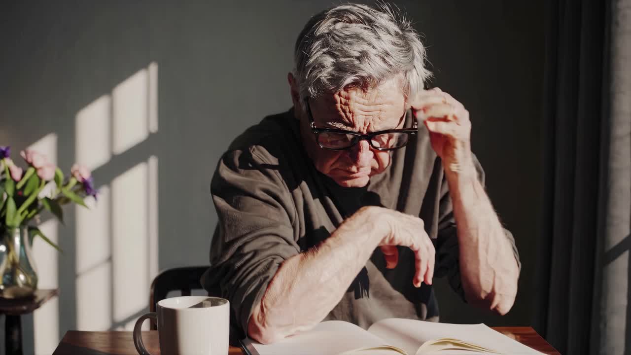 Elderly man reading at a table, hands on head, in soft natural light
