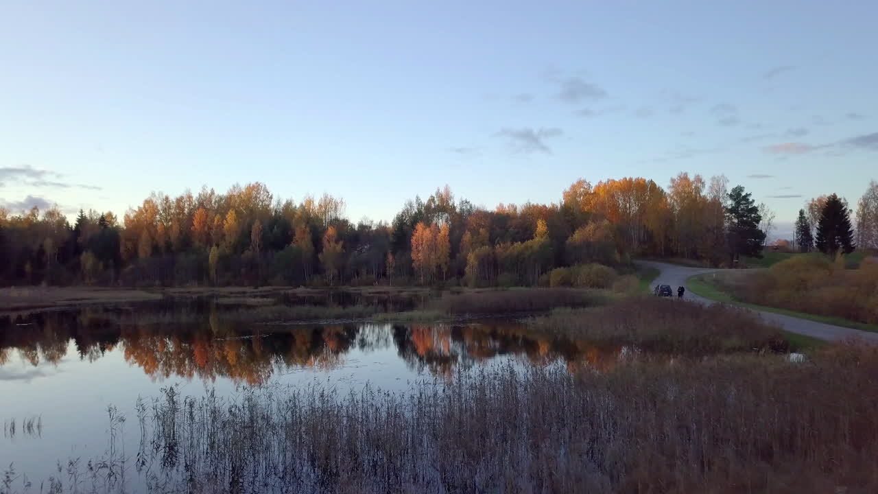 밀레니엄 세대의 남자가 가을 저녁에 연못 위로 드론을 조종하며 길 위의 차 옆에 서 있다