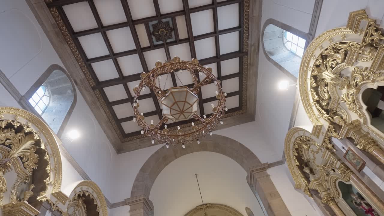 Vista di un lampadario che pende da un soffitto a cassettoni in legno all'interno di una chiesa decorata con intagli d'altare ornati, finestre e archi.