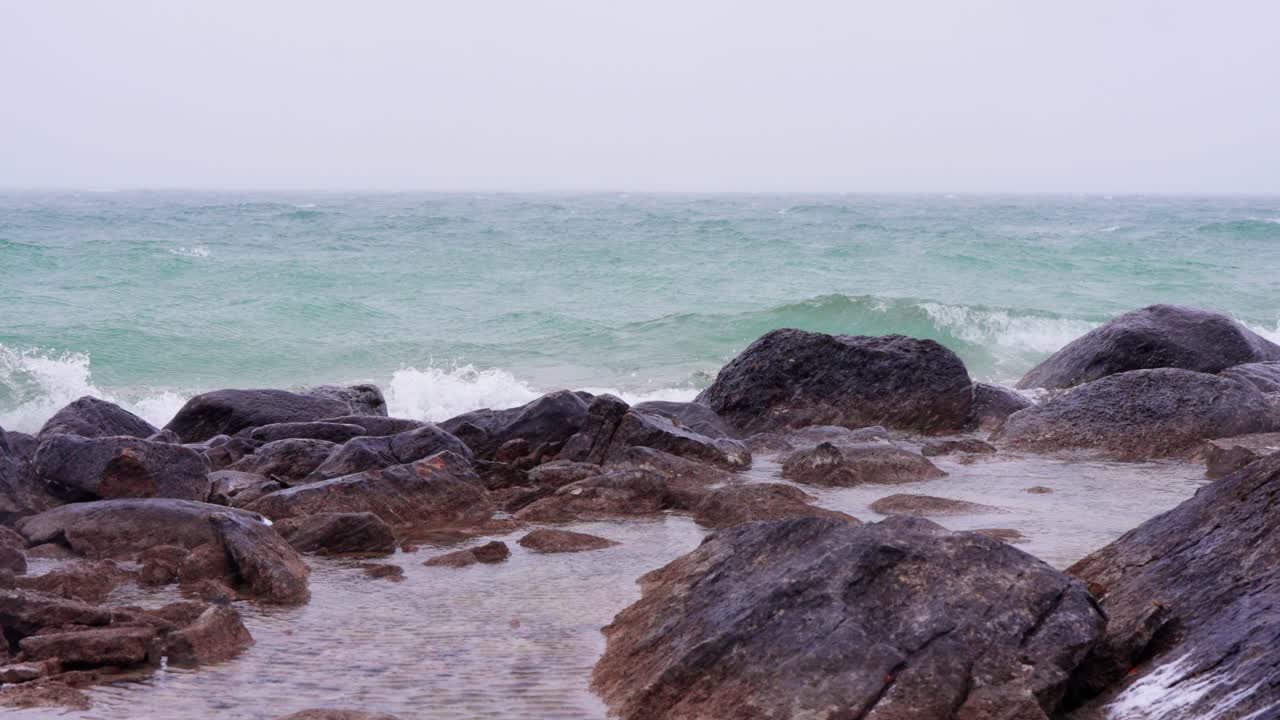 A slow motion storm reveals bright waves and brown rocks along a snowy shore