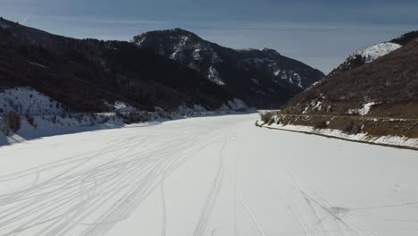 Drone-flying-sideway-to-the-left-across-a-frozen-lake-with-tracks-in-the-snow,-and-rolling-mountain-hills-on-each-side