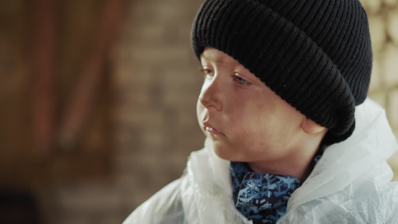 Side close up of young boy in black knit hat and protective white jacket gazing thoughtfully to side with visible dirt on face, suggesting contemplation, struggle or alert focus in harsh environment