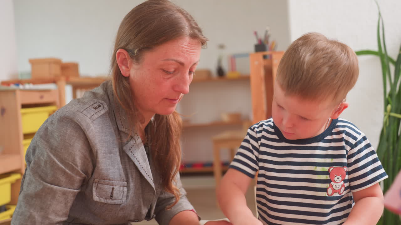 Focused teacher supports attentive little boy with hearing aid as they engage in educational task together at wooden desk in warm classroom, fostering emotional connection