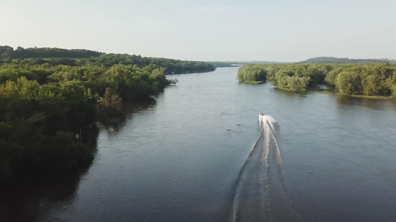 A single speedboat cuts swiftly through a broad, still river framed by lush trees on both banks, leaving a long, white wake behind as it moves downstream.