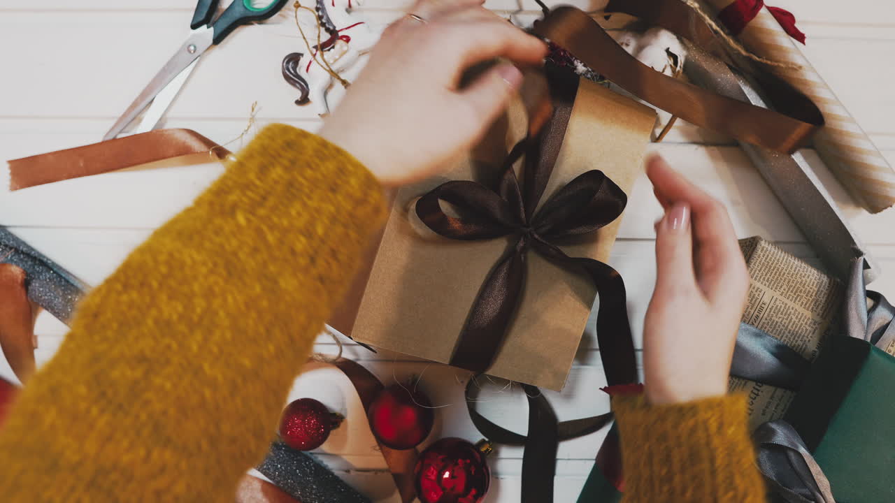 Aerial footage of woman tying red ribbon on gift. Lockdown shot of female packing present at wooden table. Overhead flat lay of art and craft equipment surrounding lady's hands.