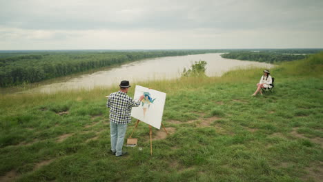 a painter in a hat, checkered shirt, and jeans, is focused on creating a masterpiece on a canvas in a grassy field beside a serene lake. a woman in a hat and white dress sits comfortably on a chair