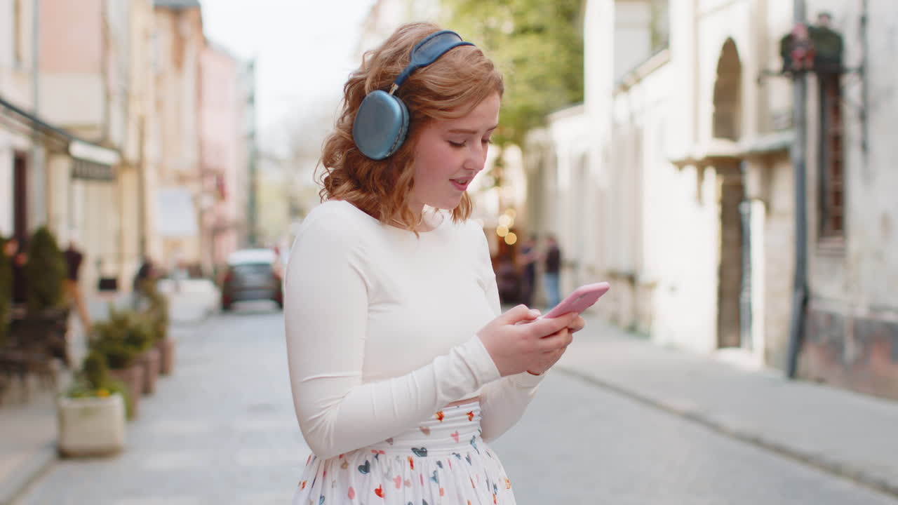 Giovane donna felice con cuffie wireless che sceglie, ascolta musica sullo smartphone e balla all'aperto