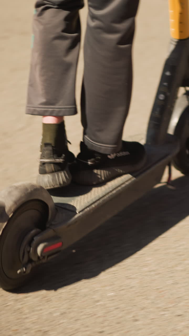 white male riding electric scooter along paved path beside green verge, denim jacket and one foot kick for balance, rear view of smooth asphalt, sunlight casting shadow, relaxed urban commute