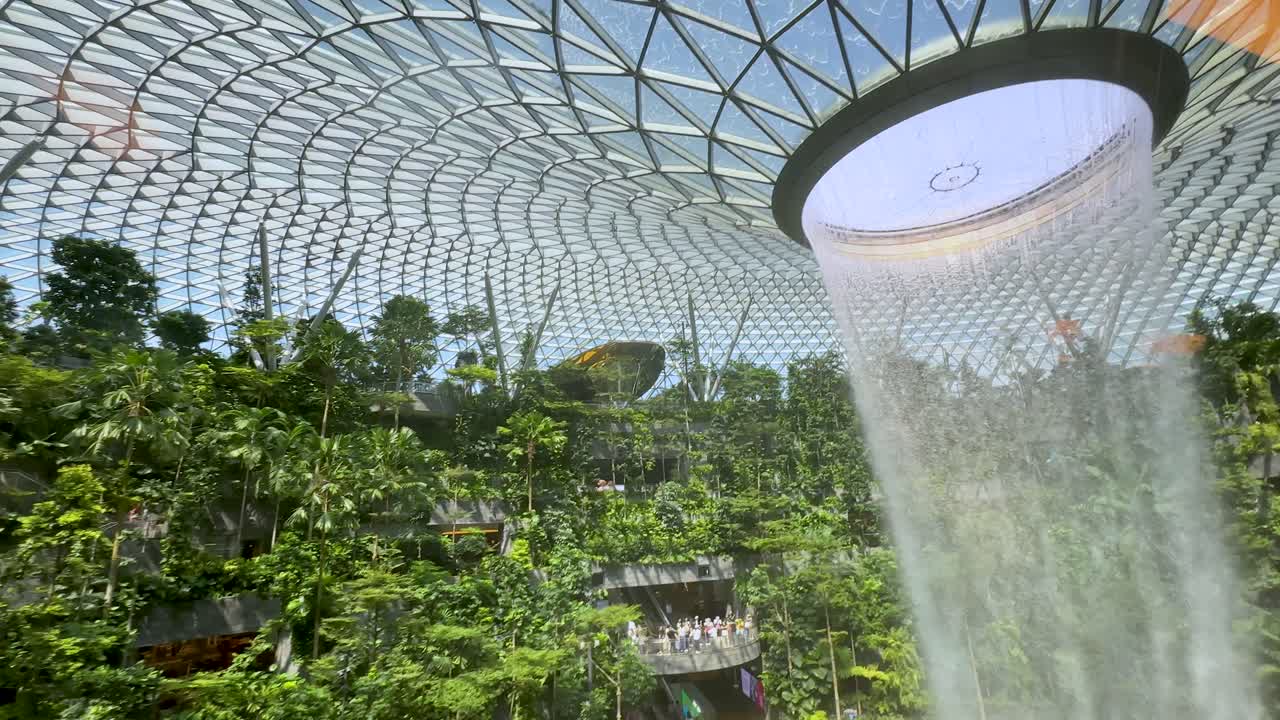 A dramatic indoor waterfall pours from a circular skylight into a verdant garden, with natural daylight illuminating the expansive glass-and-steel dome. Camera tilts downward