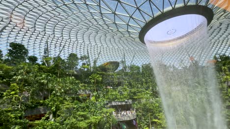 a dramatic indoor waterfall pours from a circular skylight into a verdant garden, with natural daylight illuminating the expansive glass-and-steel dome. camera tilts downward