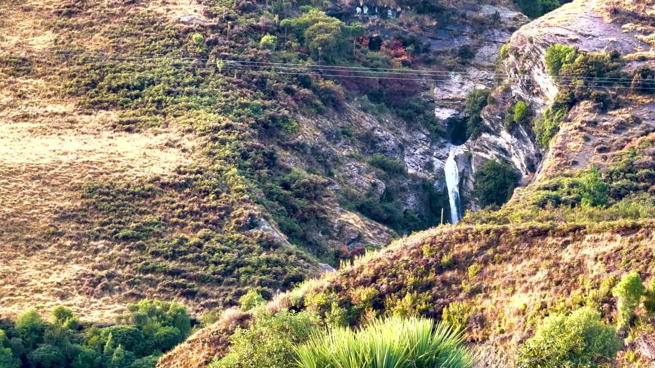A tranquil waterfall cascades down a rocky hillside in Queenstown, New Zealand, surrounded by lush greenery and bathed in soft sunlight