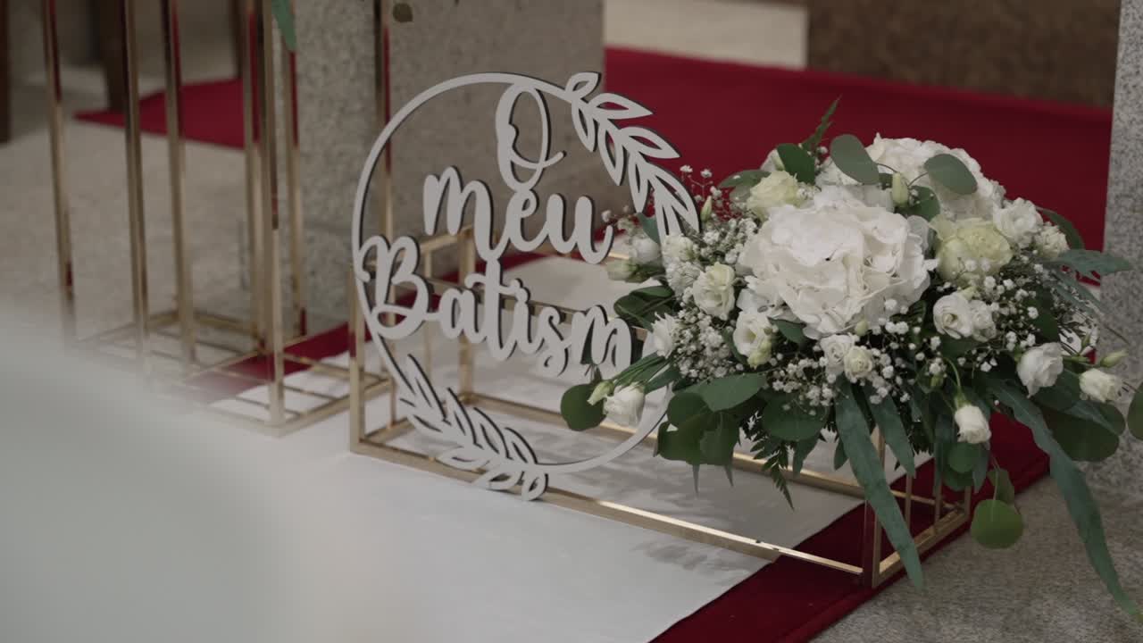 Cartello bianco per battesimo con bouquet di fiori su tappeto rosso in chiesa.