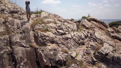 aerial: a drone rises its alttitude and reveals a girl standing at steep rocks and stones looking around the tropical landscsape, at some point the camera slowly pans to the left