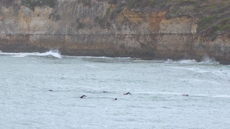 Surfers navigate waves near rocky cliffs at Port Campbell. Overcast lighting and steady camera capture the dynamic ocean scene