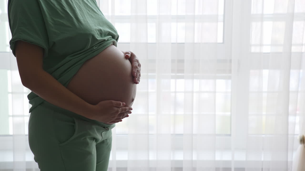 Future mom standing at the window and touching her belly gently. Expecting woman in comfortable clothes strokes her tummy lovingly.