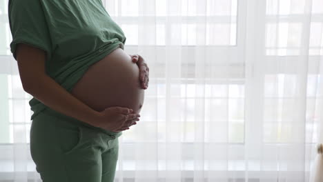future mom standing at the window and touching her belly gently. expecting woman in comfortable clothes strokes her tummy lovingly.