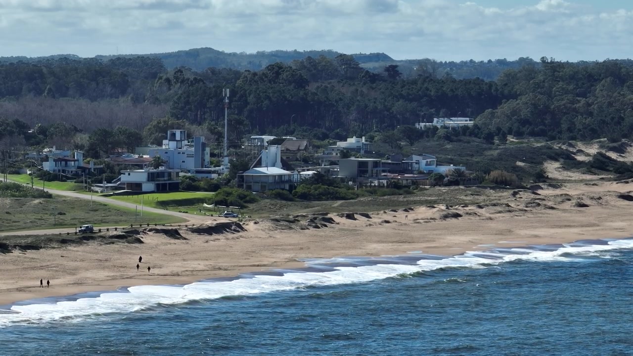 Luftaufnahme der Küste von Punta Ballena über den Sandstrand Playa Cantamar mit brechenden Meereswellen, gesäumt von Küstenhäusern und bewaldeten Hügeln in Uruguay