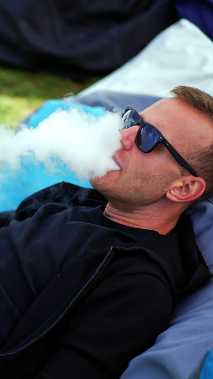 Chilling young man lying in the comfortable bean bag chair. Male in dark glasses smoking shisha and watching video on laptop. Vertical video
