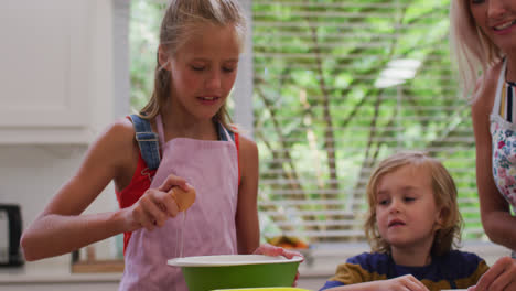 Feliz-Madre-Caucásica-En-La-Cocina-Con-Su-Hija-Y-Su-Hijo,-Usando-Delantales,-Una-Niña-Rompiendo-El-Huevo-En-Un-Tazón