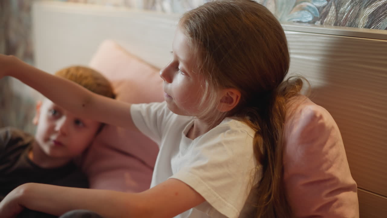 young girl in white shirt stretches arms picking bunch of berries to eat while boy lies down nearby watching her action tender sibling moment on soft pink bed against colorful decorative wall indoors
