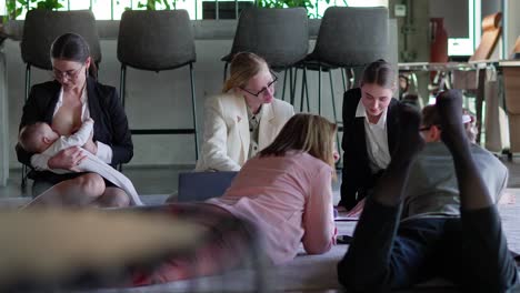 Side-view-of-a-group-of-confident-businesswomen-in-business-clothes-sitting-on-a-soft-carpet-in-the-office-and-communicating-with-each-other-while-their-colleague-a-brunette-girl-in-business-clothes-breastfeeds-her-infant-child-in-the-office