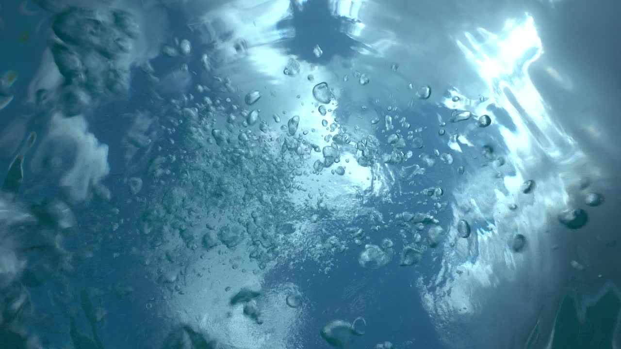 Camera points towards the surface of the ocean capturing the surface and a scuba diver's bubbles