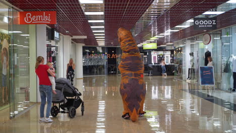 man in orange costume of dinosaur walking away from camera. animator visiting the big supermarket surprising shop customers.