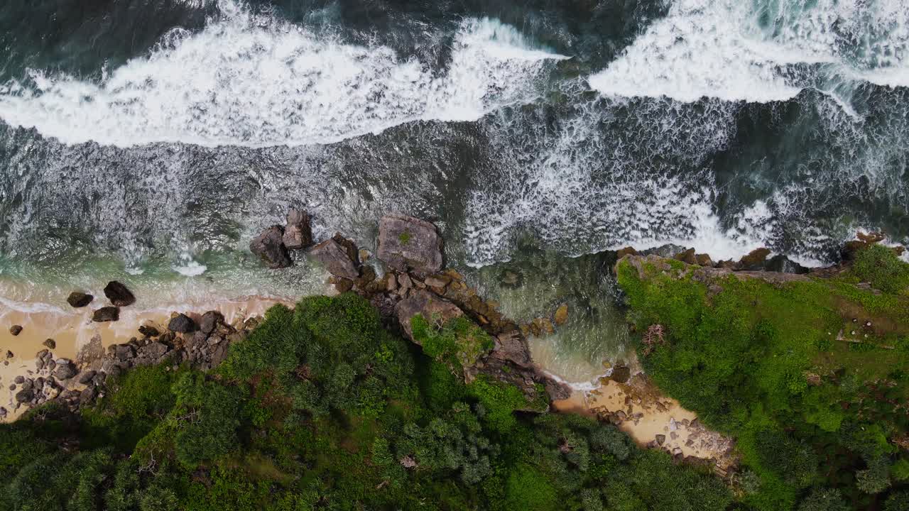 Sliding overhead drone shot of cliff on the beach overgrown with green grass and trees and big sea wave - Tropical Beach, Indonesia