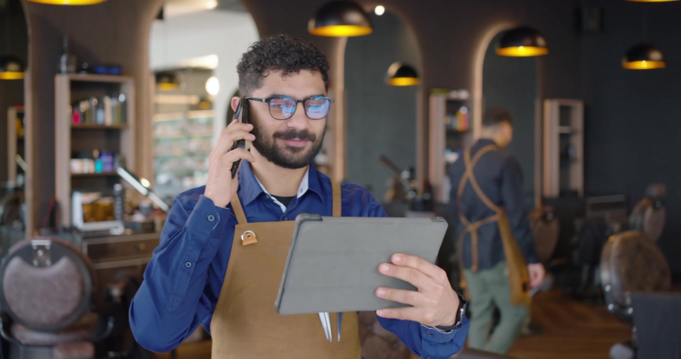 Barbeiro afro-americano atendendo o telefone enquanto lê informações em tablet na barbearia, usando avental de couro