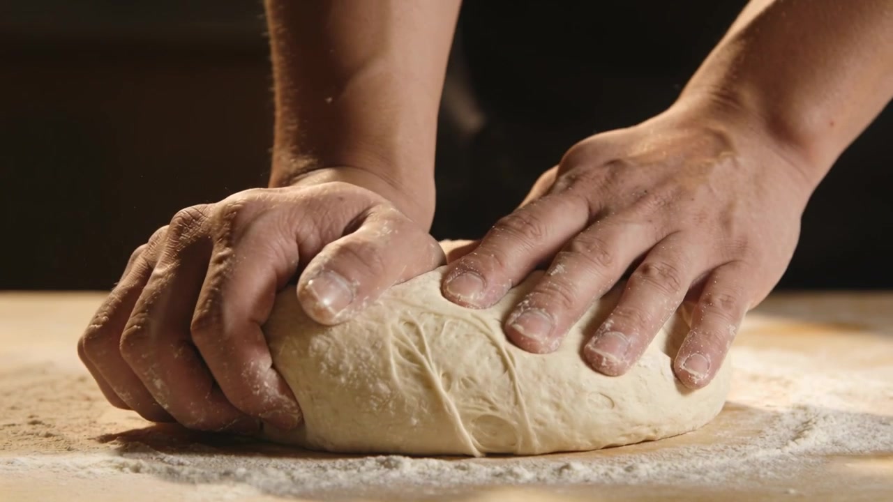 Lowering hands with bare forearms pressing and folding dough on floured wooden bench, making bread