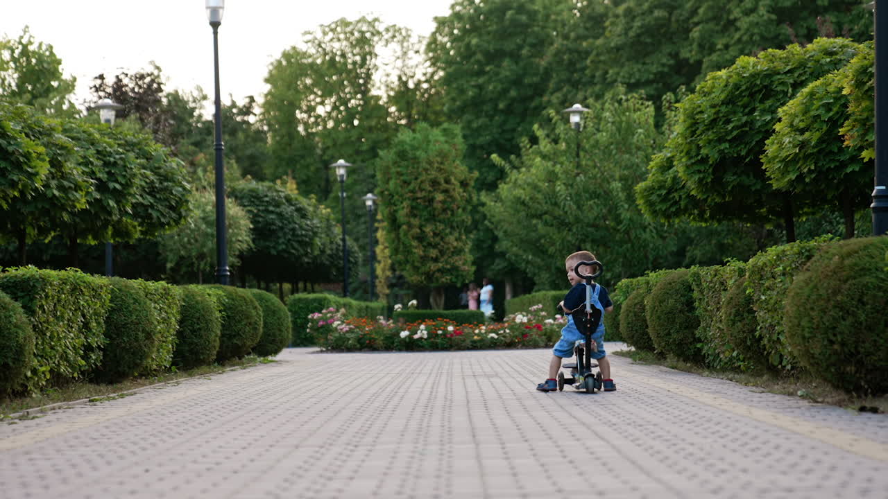 Rear view of a baby boy sitting on a bike. Kid waves his head and then turns to camera. Park in summer at backdrop.