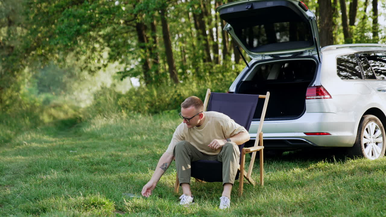 Relaxed man sitting in folding chair puts on glasses. Freelancer takes his laptop, opens and starts work. Nature backdrop.