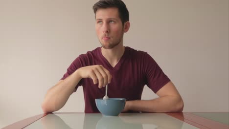 Man-sitting-at-table-eating-his-cereals-and-twisting-with-spoon-while-looking-in-the-distance,-still-shot-indoors