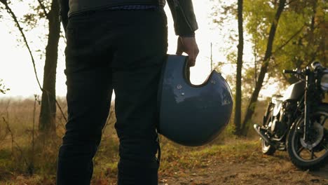 back view of a man in black jeans and leather jacket holding his helmet in his hand while standing by his cool bike on a sunny