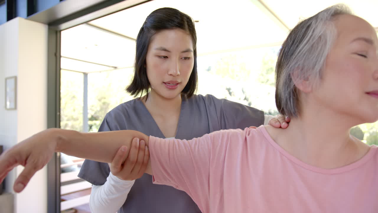 Physiotherapist assisting senior asian woman with arm exercises at home therapy session