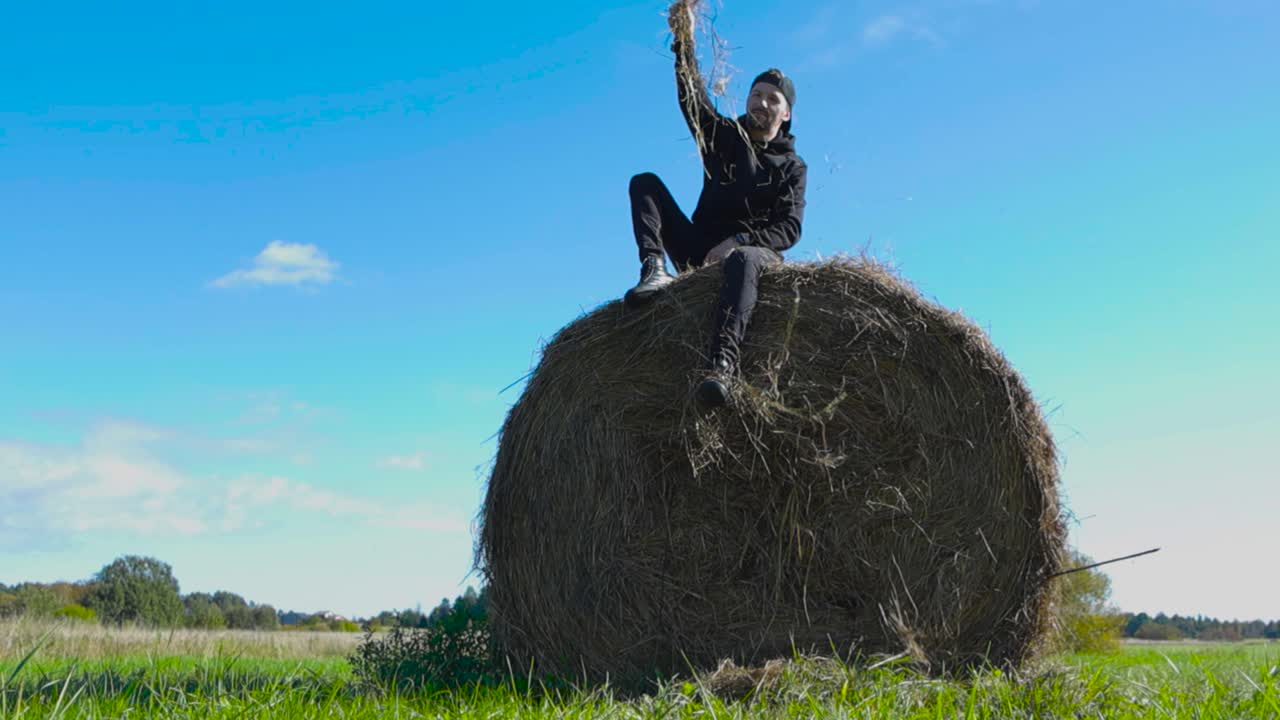 Caucasian man with black clothes sitting on top of a hay bale in a farm field and drops hay to the ground in slow motion from his hands. Summer time and sun is shining with blue sky in the background.