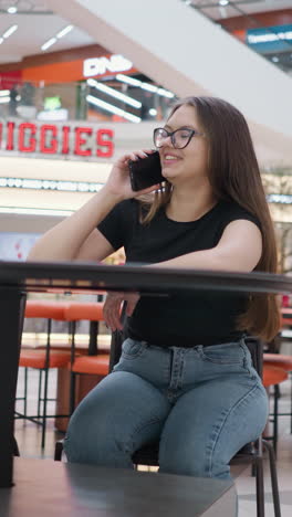 joven sentada en una mesa en el centro comercial, felizmente haciendo una llamada telefónica con bolsas de compras a su lado, otras personas están sentadas a su alrededor, con el letrero de biggies visible en el fondo, creando una atmósfera animada