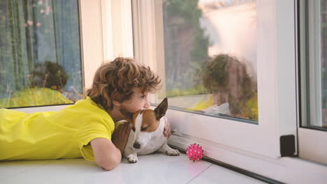 Blonder-Junge-Mit-Lockigen-Haaren,-Der-Mit-Seinem-Hund-Auf-Dem-Boden-Liegt,-Ihn-Streichelt-Und-Aus-Dem-Fenster-Schaut-1