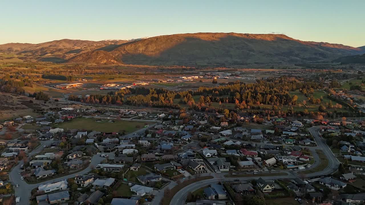 Drone Hyperlapse flight over suburb city with traffic on Main Street in wanaka town, New Zealand. Lighting golden peaks at sunrise in distance