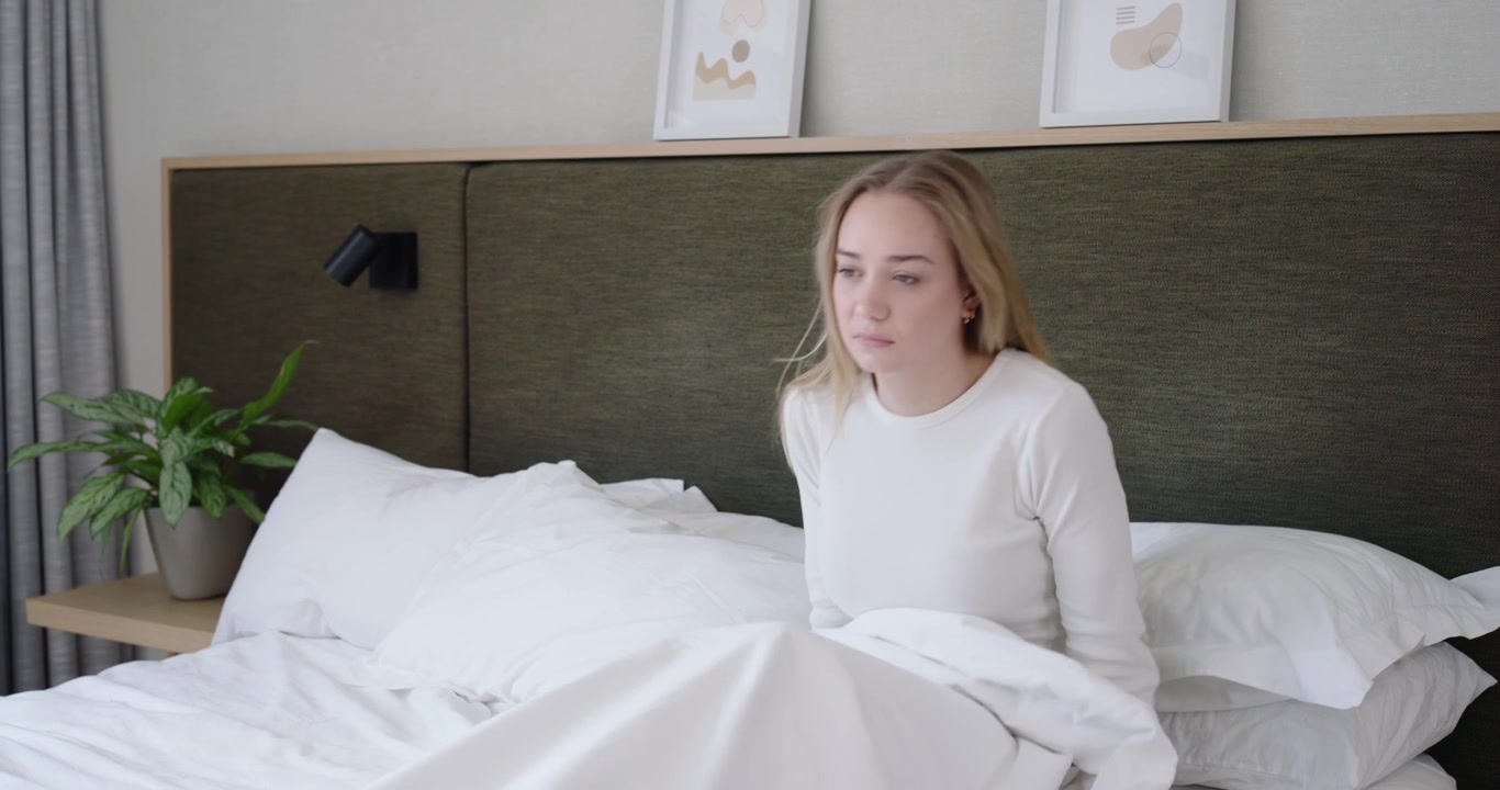 Woman waking as left curtain light entering, propping up while rubbing temples, in bed