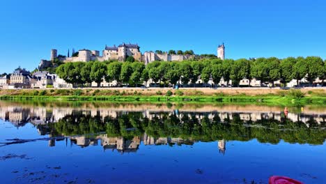view of chinon fortress with trees and medieval walls mirrored in the calm waters of the vienne river under a clear blue sky - france