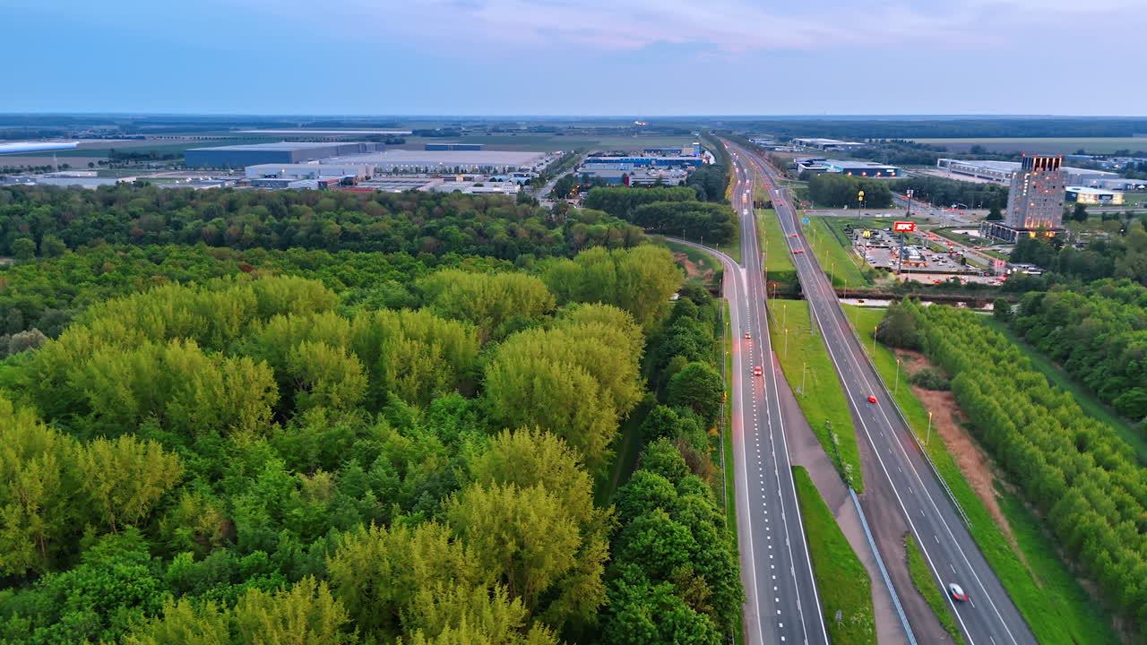 Cars going quickly by the highway passing near the green deep woods. Infrastructure of the modern little town at backdrop. Aerial view.