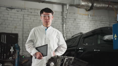 mechanical engineer wearing lab coat and protective glasses stands in automotive workshop with hand on waist while holding tablet and nodding, background features parked car and work tools